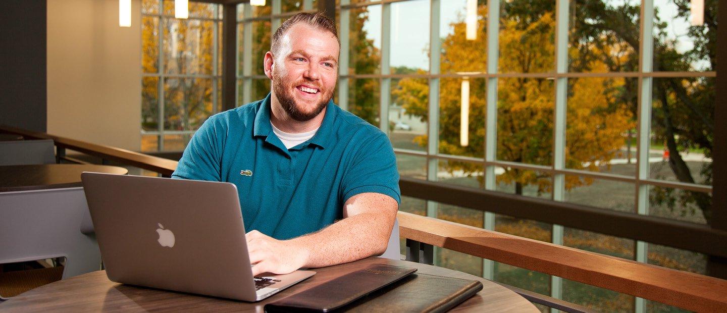 A man seated at a table with a laptop and a view of trees with yellow leaves through windows behind him.